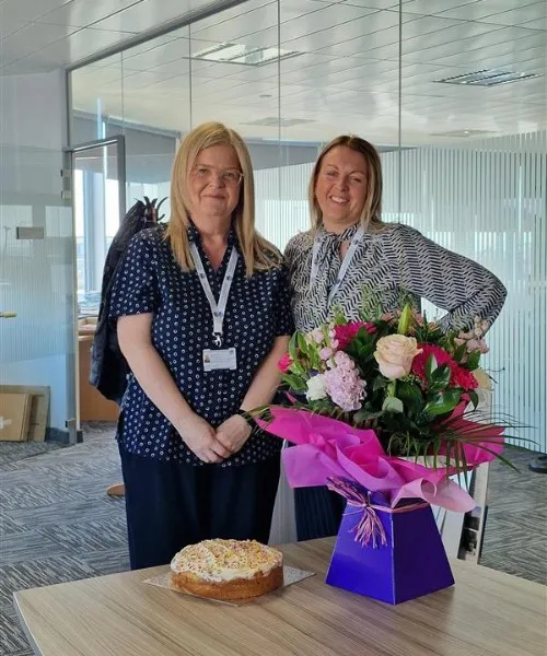 Two women in an office with a bunch of flowers and a cake.